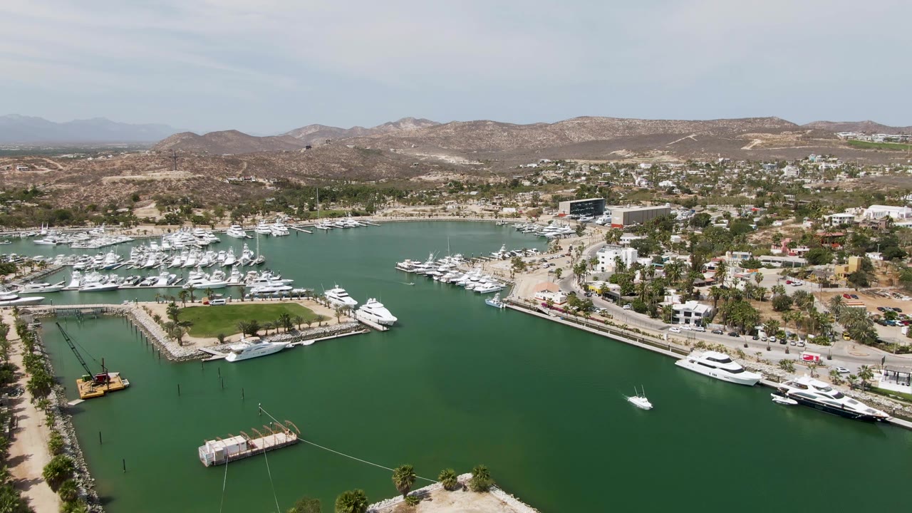 Opening aerial frame over Puerto Los Cabos Beach marina with green water in front, docked yachts along piers, coastal buildings, dry hills, pale sky, and no visible people.