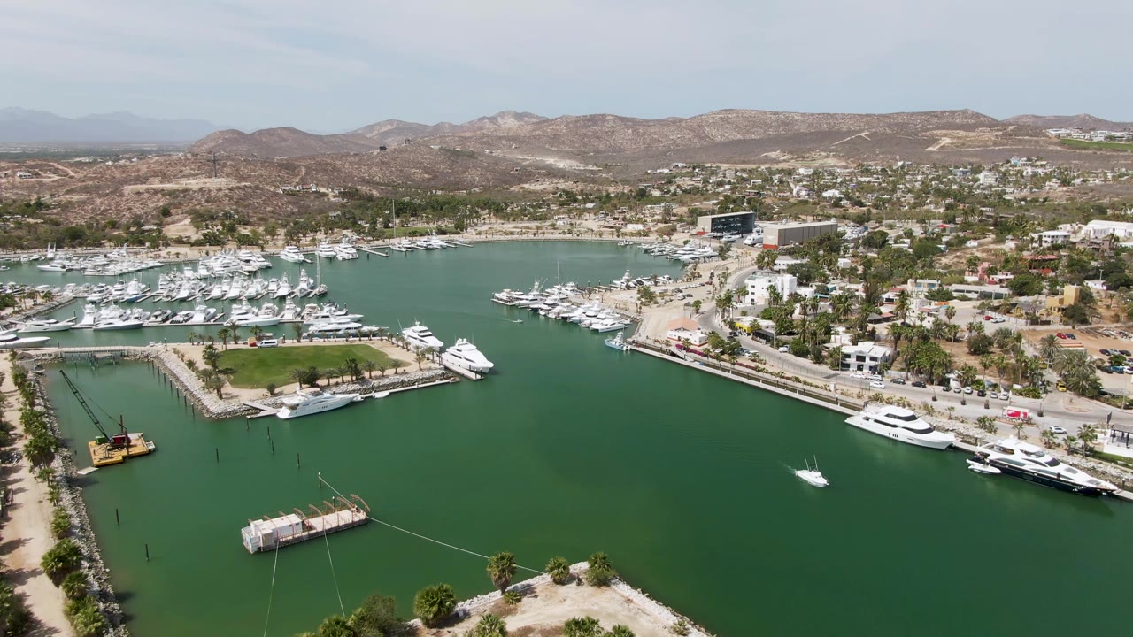 Mid-clip aerial frame over Puerto Los Cabos Beach with the marina basin centered, white yachts lining the docks, a small craft on the water, arid hills behind town, and no visible people.