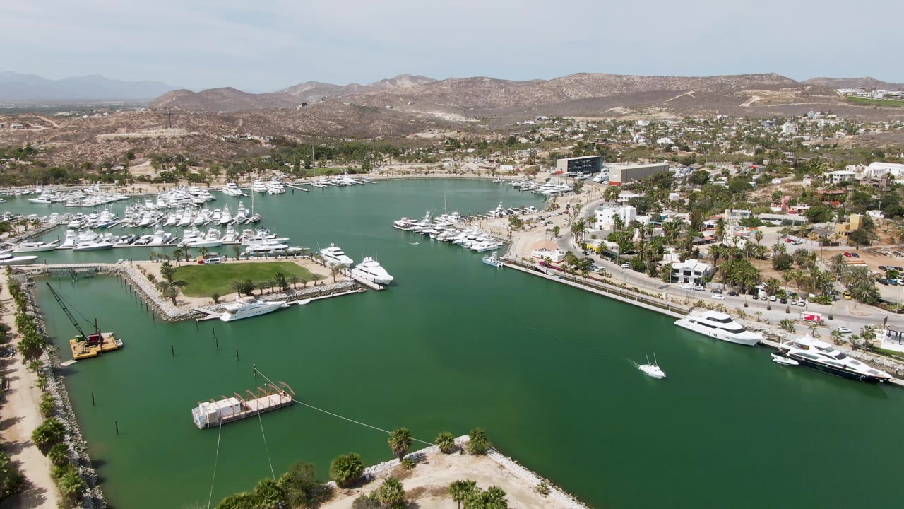 Aerial view of a marina entrance area with green water, piers, yachts, and coastal hills.