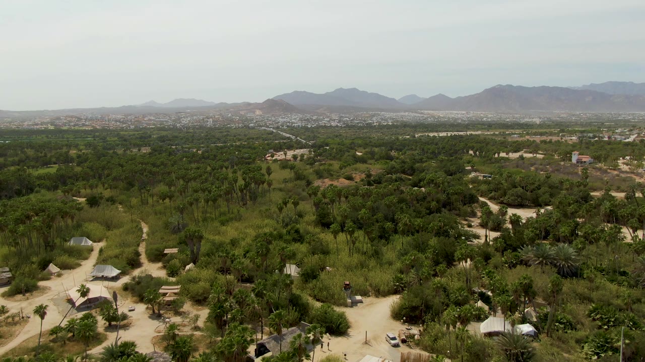 Mid-clip aerial frame in Baja California Sur shows sandy tracks winding between green trees and scattered huts, with hazy mountains across the horizon and no visible people.