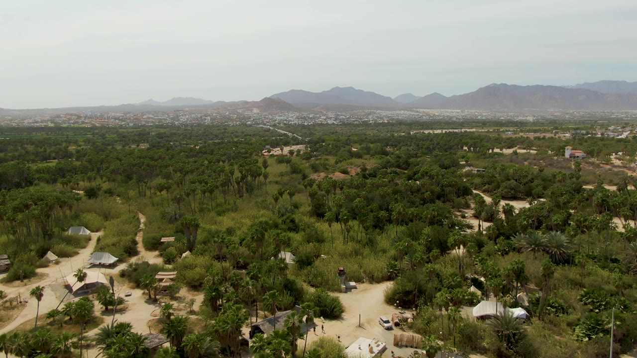 Aerial view across green rural land with huts, dirt paths, distant buildings, and mountains.