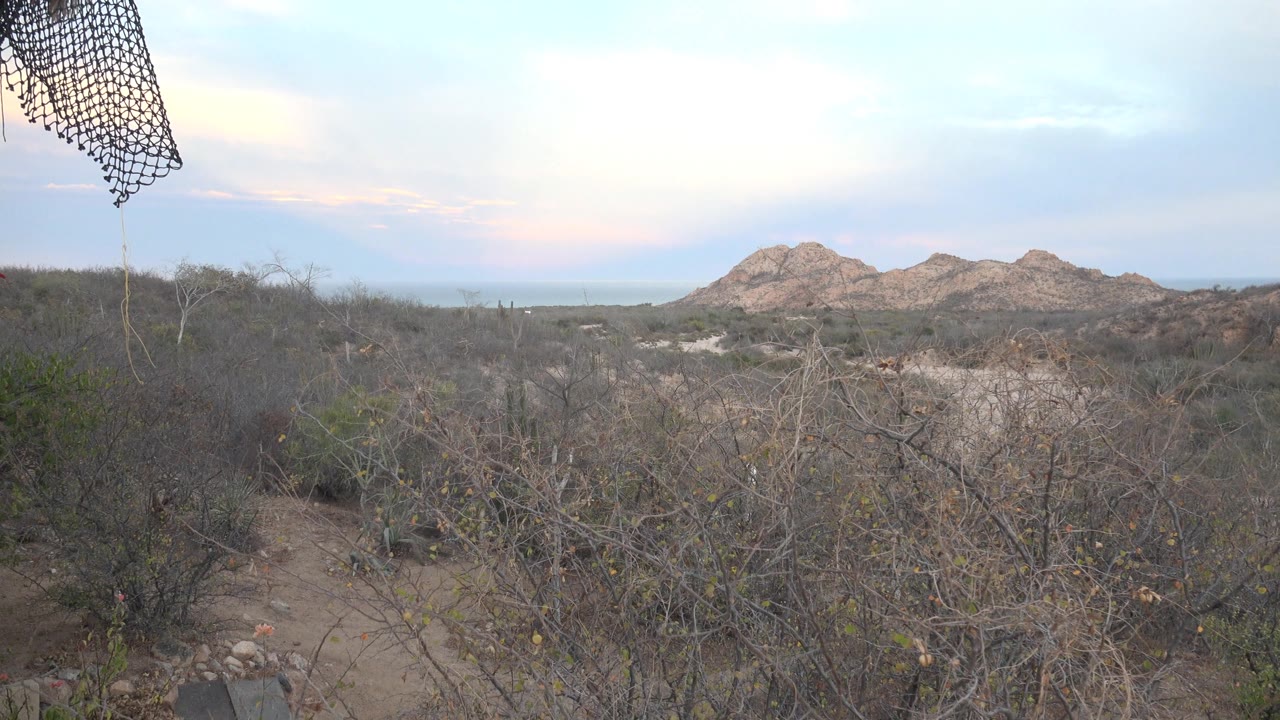 Balcony netting hangs at the upper left over dry brush, dirt paths, and rocky hills near Cabo Pulmo, Baja California Sur, with no visible people.