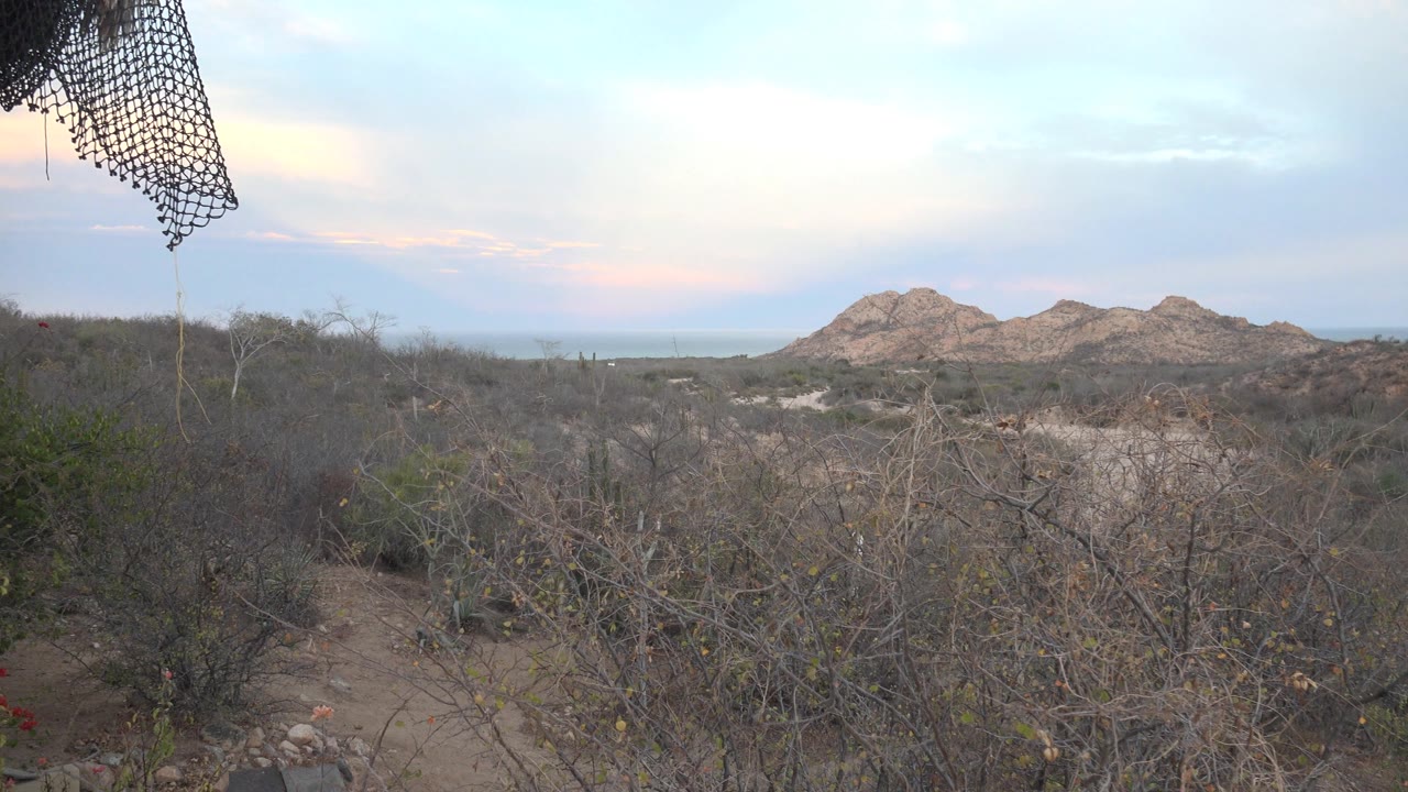 A wider balcony-framed view shows dark netting at the upper left, scrubby desert foreground, and rocky hills under pastel clouds in Cabo Pulmo, with no visible people.