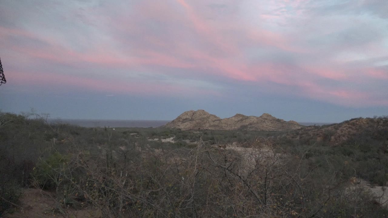 Open Cabo Pulmo desert scrub fills the foreground with rocky hills beyond, a wide blue-gray sky, pink cloud bands, and no visible people.