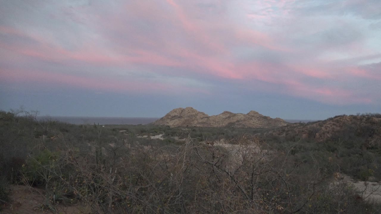 A Cabo Pulmo desert horizon shows low rocky hills, dark scrub in the foreground, streaked pink dusk clouds, and no visible people.