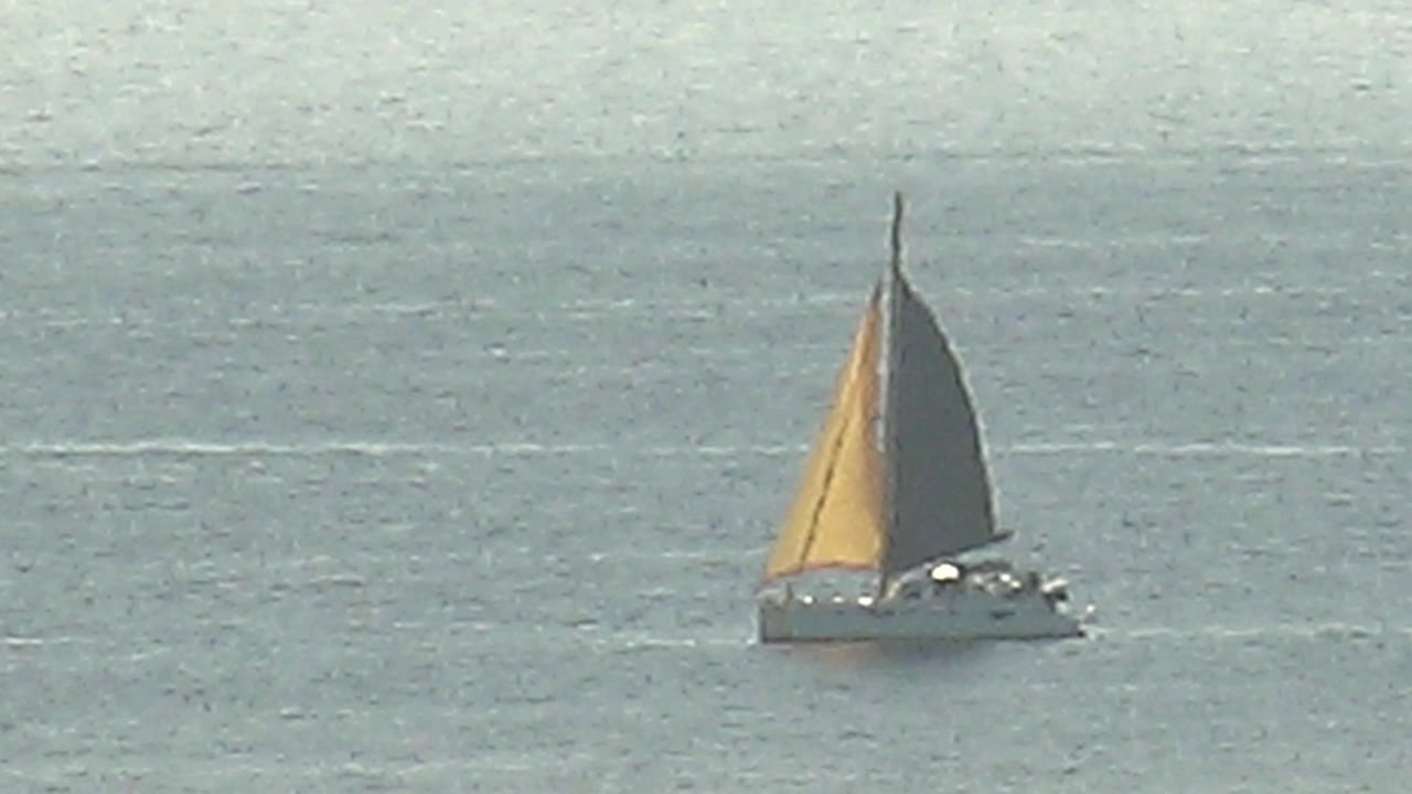 A distant gray sailboat with yellow and dark sails moves across calm blue-gray water in Cabo Pulmo, Baja California Sur, with no visible people.