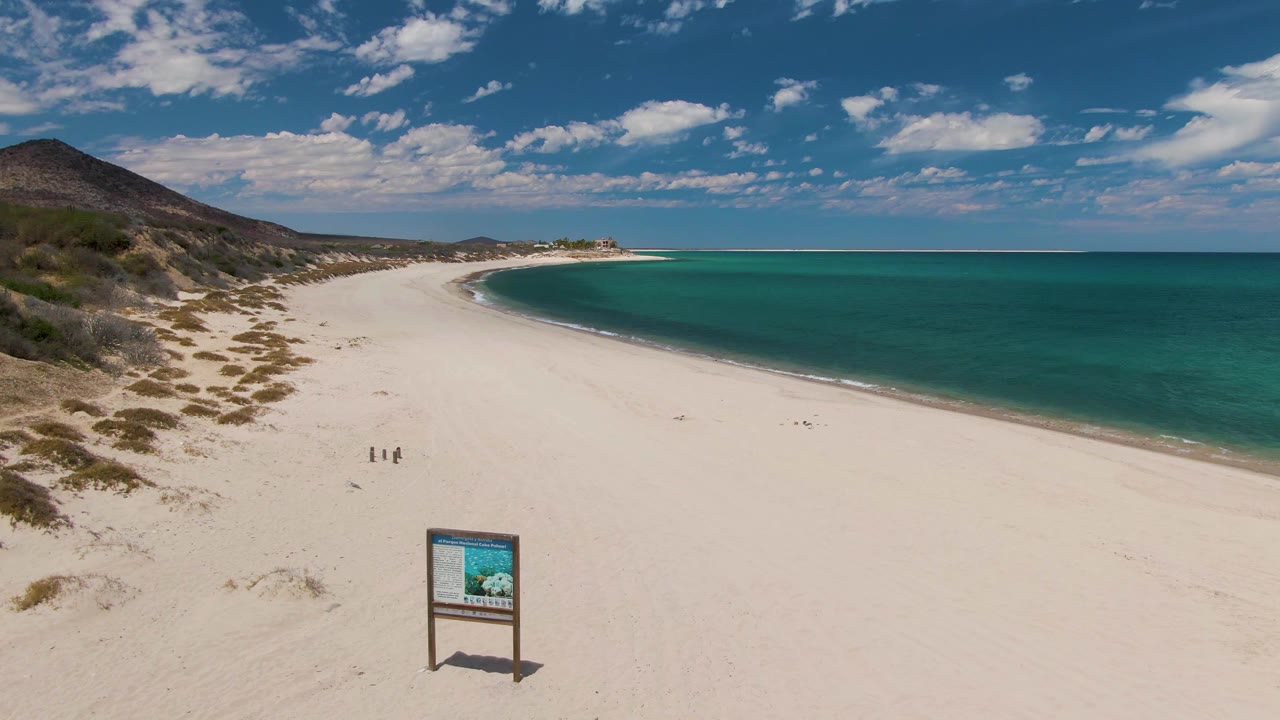 Aerial opening frame at Barakas Beach, Baja California Sur, showing white sand, turquoise ocean, a foreground beach sign, low dunes, hills, clouds, and two distant people walking near the center of the beach.