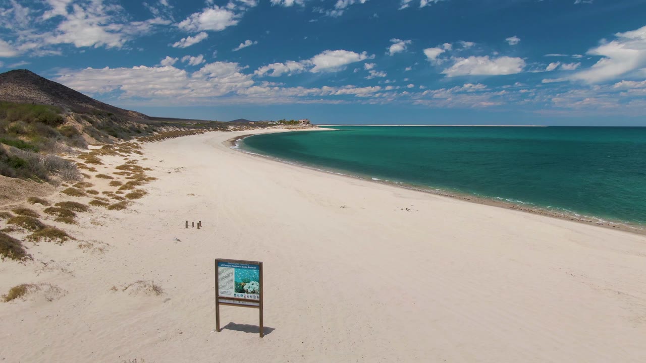 Aerial view at Barakas Beach showing the beach sign near the lower foreground, two tiny people on the open sand, pale dunes along the left side, and calm turquoise ocean under a clouded blue sky.