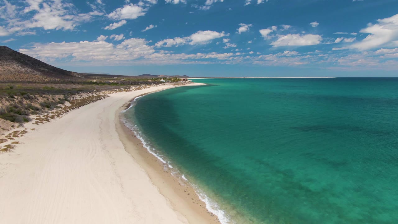 Drone view at Barakas Beach with the sandy shoreline running diagonally through frame, calm turquoise water to the right, dry scrubland to the left, distant buildings, and no visible people.