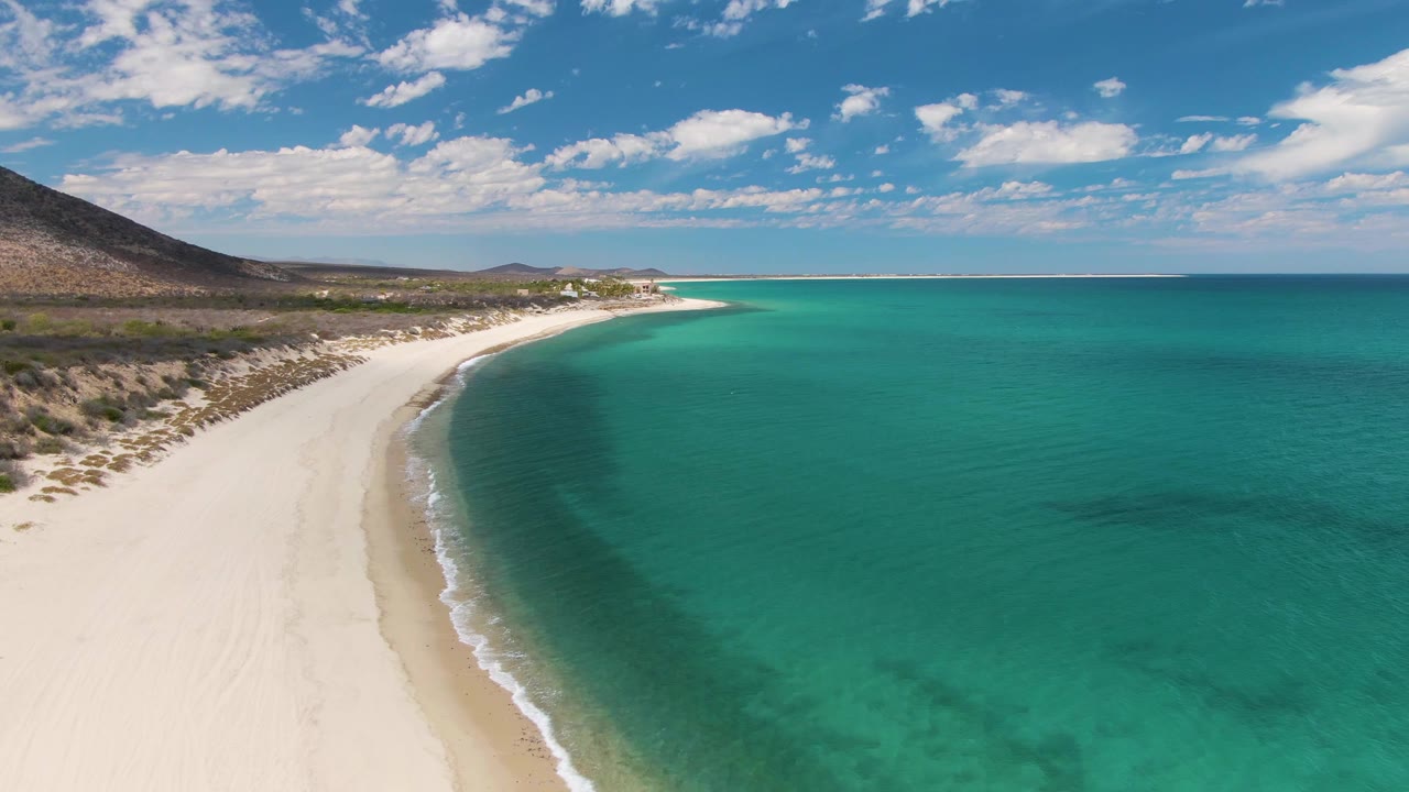 Barakas Beach aerial frame showing pale sand curving below a rocky hillside, transparent turquoise shallows, a darker patch in the water, scattered clouds, and no visible people.