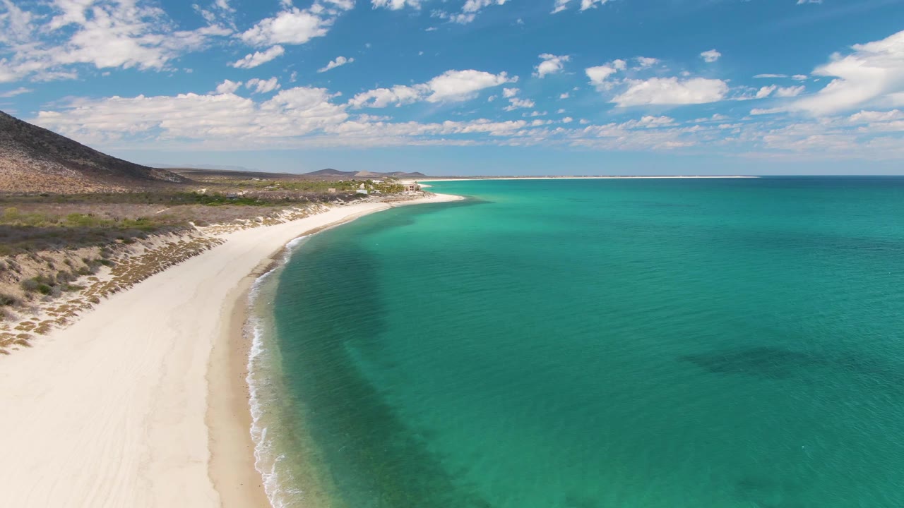 Aerial view over Barakas Beach with broad turquoise water filling the right side, pale sand and low surf on the left, dry coastal terrain behind the beach, and no visible people.