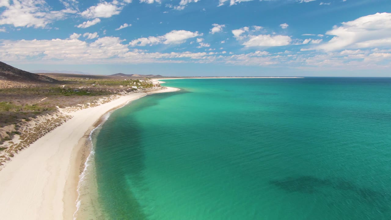Aerial frame of Barakas Beach in Baja California Sur, with white sand curving along turquoise water, sparse inland vegetation, scattered clouds, and no visible people.