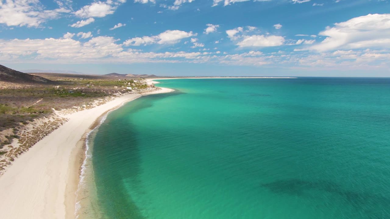 Aerial view over Barakas Beach, Baja California Sur, showing the pale shoreline slightly farther along the curve, teal shallows, inland scrub, distant hills, and no visible people.