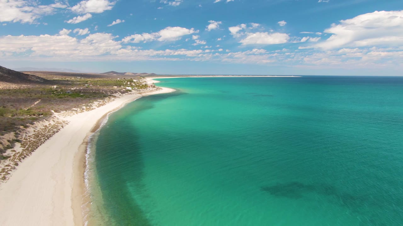 Aerial frame from Barakas Beach in Baja California Sur with the beach curve extending into the distance, broad turquoise water to the right, low land to the left, and no visible people.