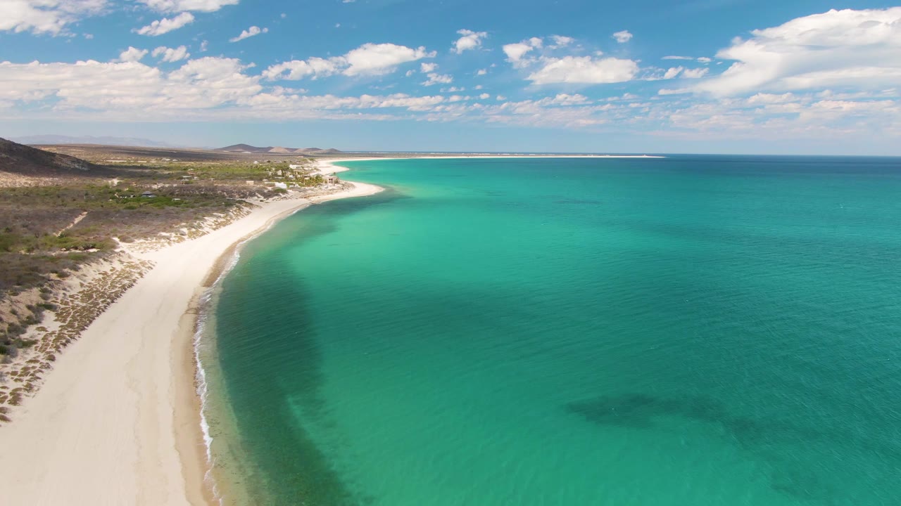 Aerial view of Barakas Beach, Baja California Sur, with more open water filling the right side, the pale beach running up the left edge, scattered clouds above, and no visible people.