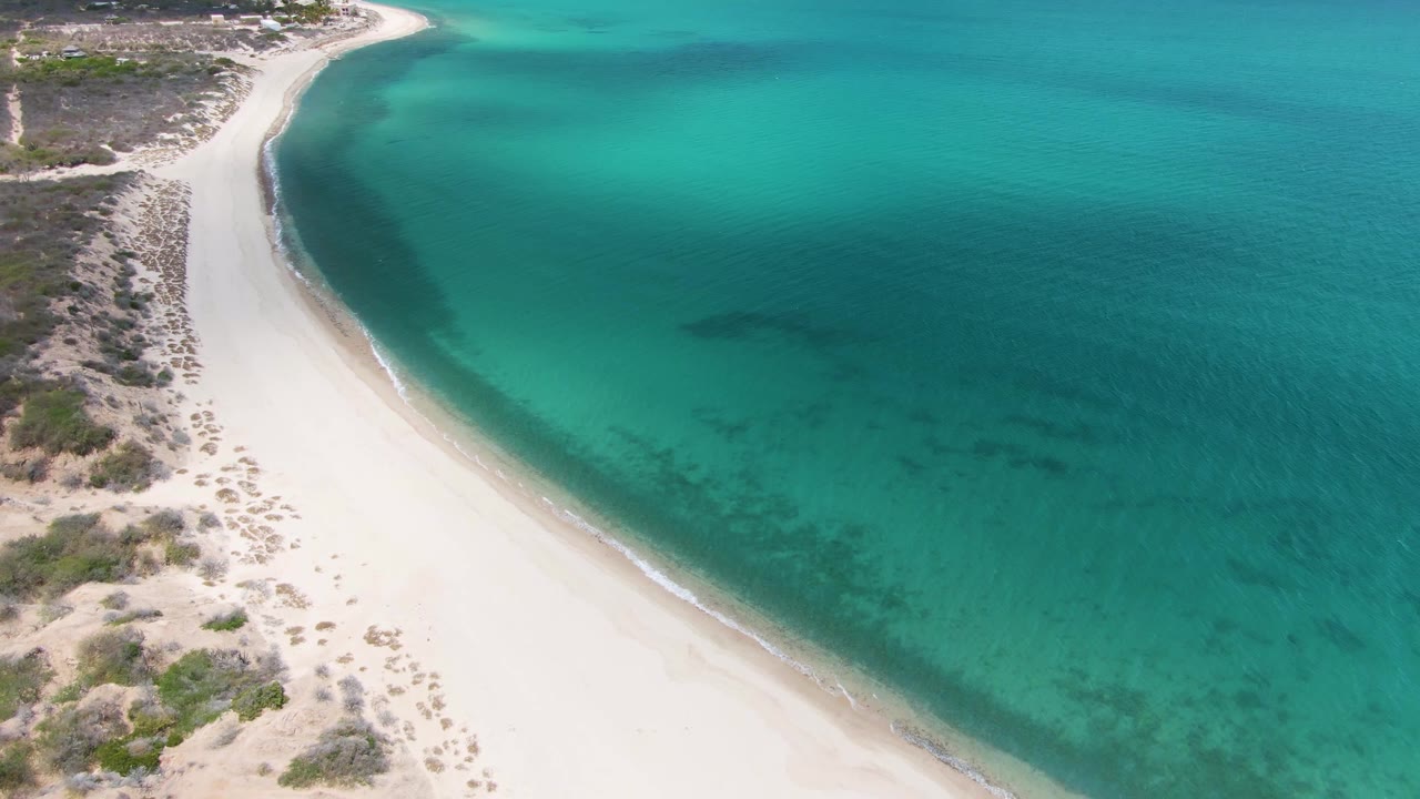Aerial frame at Barakas Beach with the shoreline running diagonally, pale sand on the left, clear turquoise shallows on the right, darker underwater areas offshore, and no visible people.