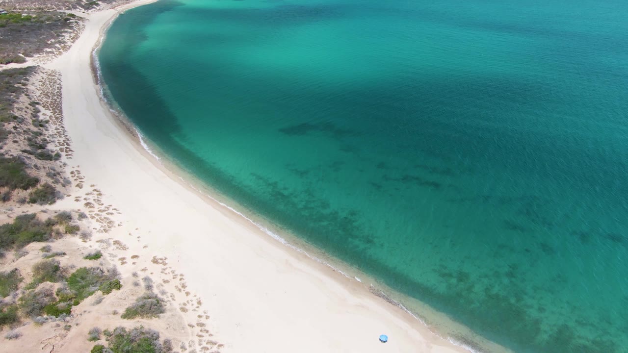 Aerial view of Barakas Beach with the sandy curve along turquoise water, scrub vegetation inland, a small blue object near the lower right beach area, and no visible people.
