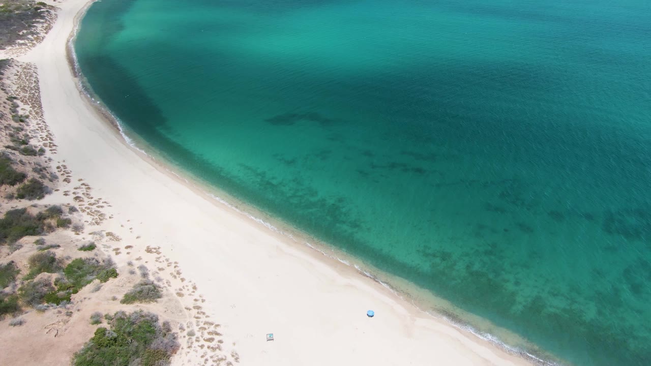 Aerial frame over Barakas Beach showing a wider sweep of white sand, calm turquoise water, faint wave edge, inland scrub, a small pale item near the lower sand, and no visible people.
