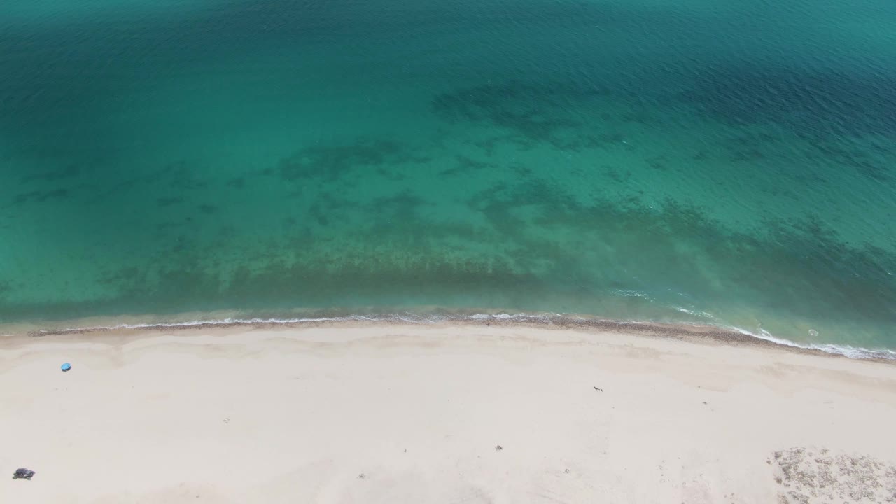 Aerial frame at Barakas Beach showing the turquoise surf line shifted slightly along pale sand, with offshore dark patches and no visible people.