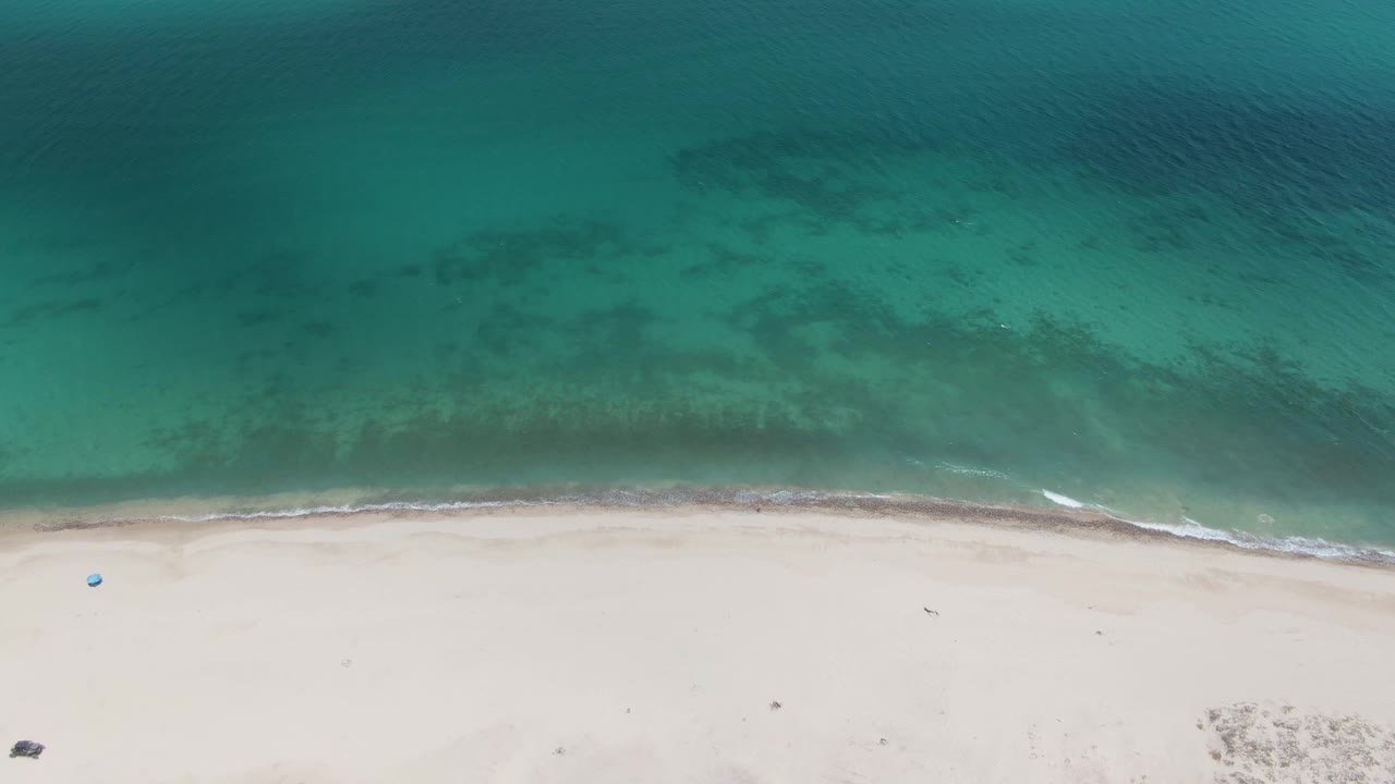Top-down view of clear green-blue water beside empty pale sand at Barakas Beach, with a narrow foam line and no visible people.