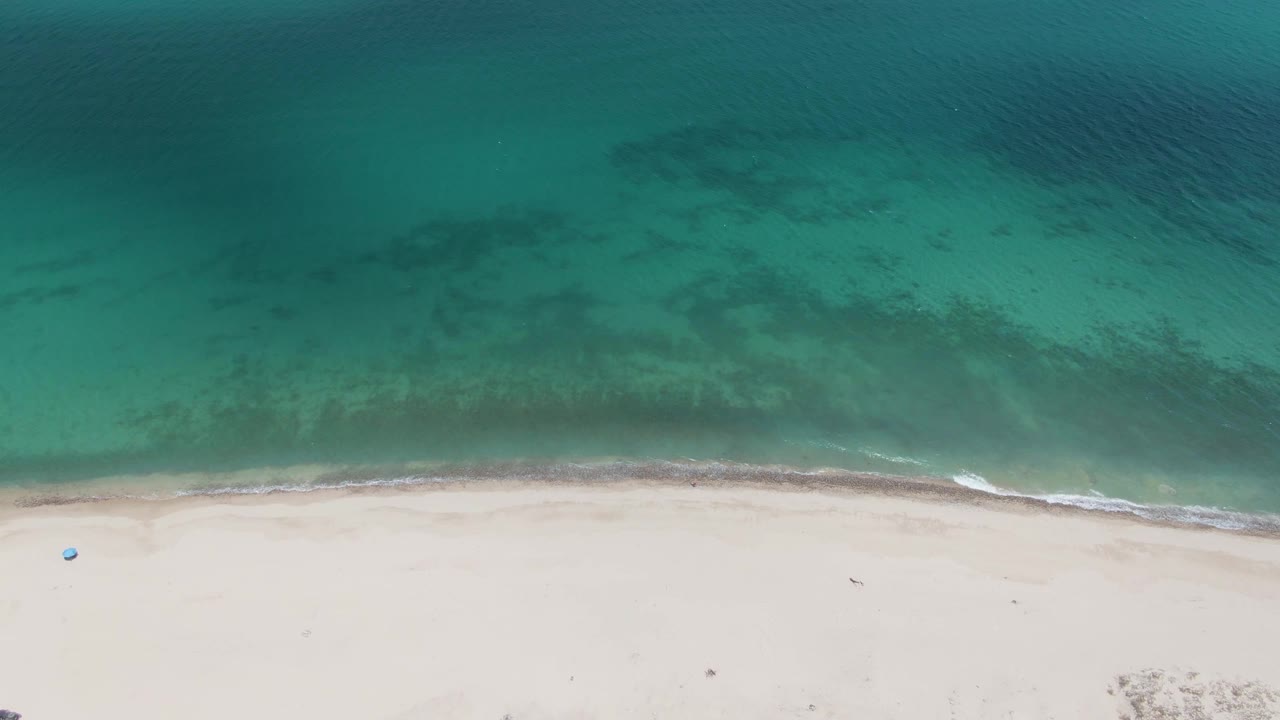 Aerial view at Barakas Beach with darker underwater patches in turquoise water and a soft wave line touching pale sand, with no visible people.