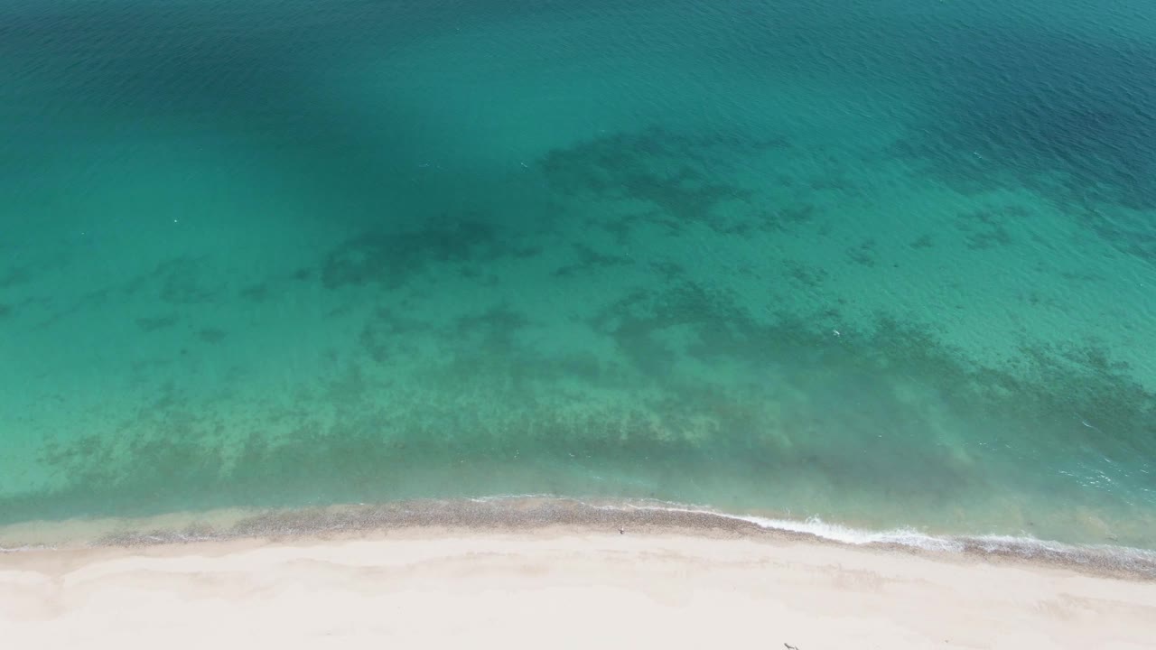 Overhead view of clear turquoise water reaching a pale sandy shoreline at Barakas Beach, Baja California Sur, with darker underwater patches and no visible people.