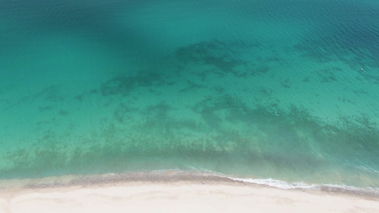 Top-down frame at Barakas Beach showing turquoise shallows, submerged dark patches, and a narrow white wave edge along the pale sand with no visible people.