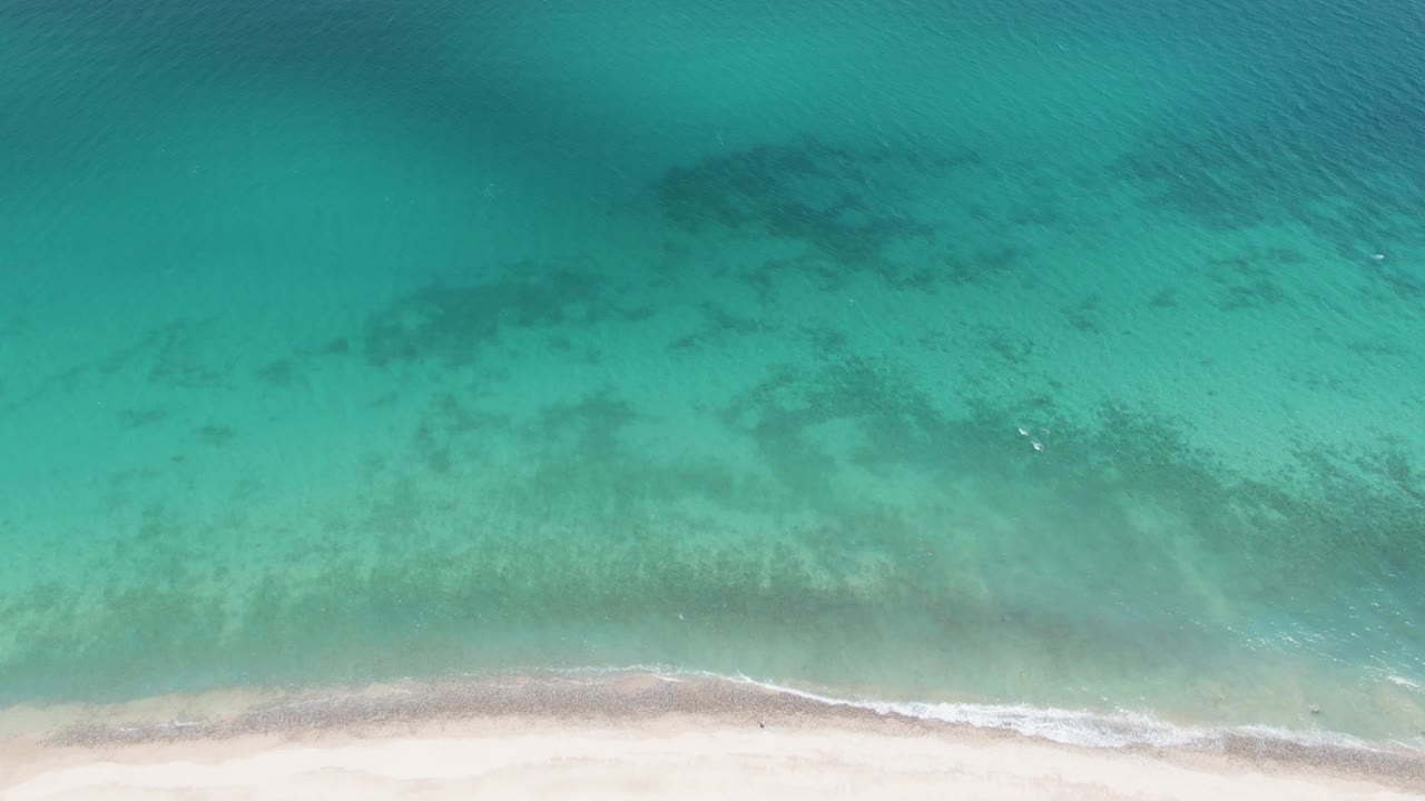 Aerial view of Barakas Beach with the sandy strip along the lower frame, turquoise water across the scene, darker seabed patches offshore, and no visible people.
