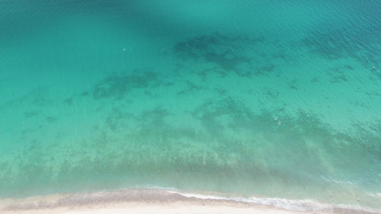 Overhead Barakas Beach frame with shallow turquoise water, broad dark areas beneath the surface, a pale beach edge at the bottom, and no visible people.