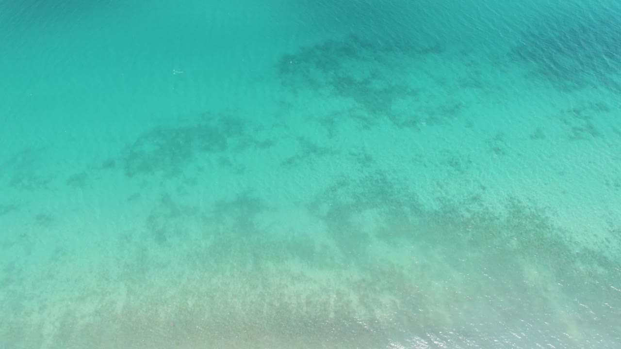 Top-down view of turquoise water at Barakas Beach, with pale seabed and darker underwater patches visible beneath rippled surface, no visible people.