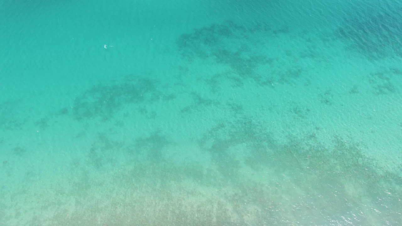 Clear aqua water at Barakas Beach shows faint coral-like dark patches and pale sand below the lightly rippled surface, no visible people.