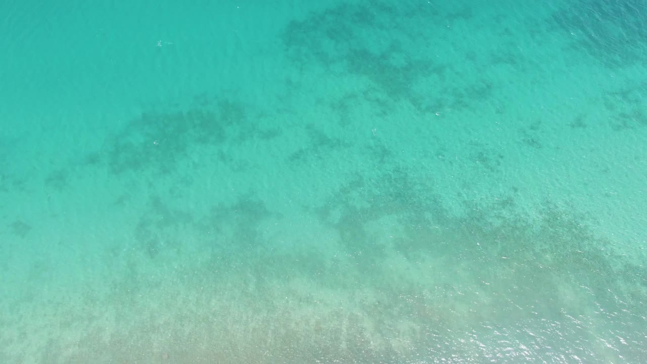 Aerial frame over Barakas Beach water with turquoise surface ripples and darker seabed shapes spread beneath the clear shallows, no visible people.