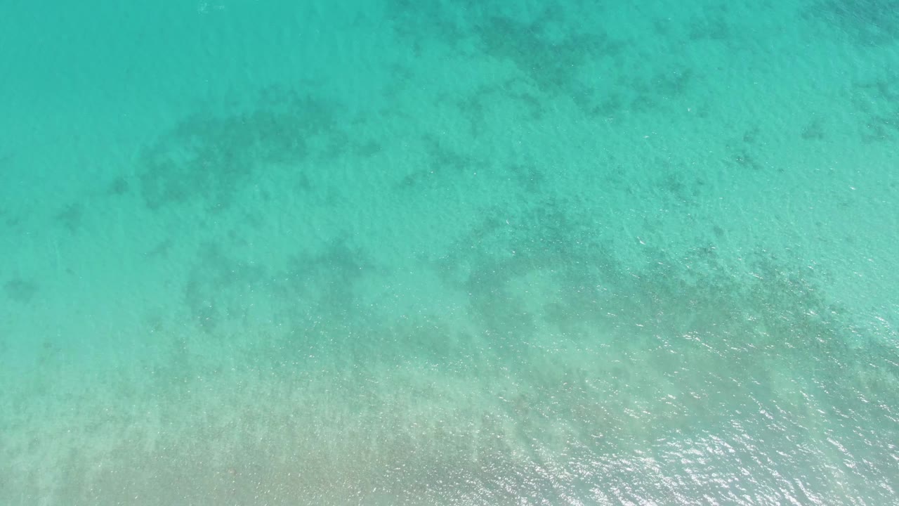 Top-down Barakas Beach water fills the frame with pale seabed tones, muted dark patches, and soft reflected light on the rippled surface, no visible people.