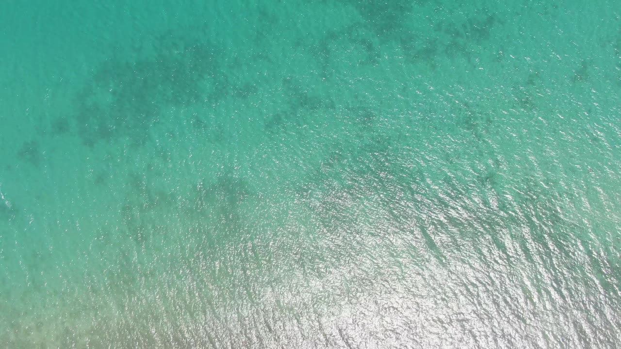 Aerial view of Barakas Beach shallow ocean with brighter sun shimmer spread across the lower part of the frame and no visible people.