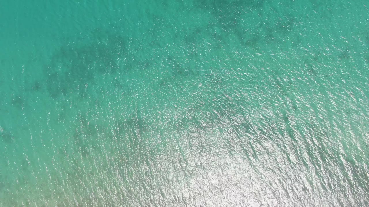 Top-down Barakas Beach water detail showing transparent turquoise sea, faint darker seabed patches, rippled texture, and no visible people.