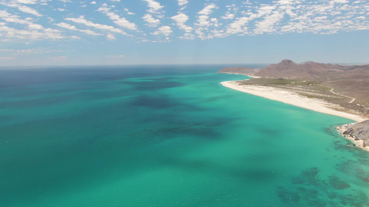 Aerial view over Barakas Beach in Baja California Sur, with turquoise water in the foreground, white sand curving at right, low brown mountains beyond, and no distinct people visible at this distance.