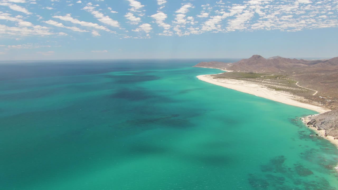 Aerial view of Barakas Beach with more open water across the left side, the white beach continuing along the right edge, mountains behind the shore, and no distinct people visible in the still.