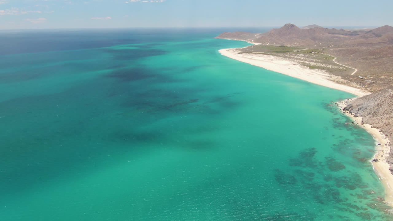 Drone frame over Barakas Beach showing broad turquoise water in the foreground, darker submerged areas offshore, a white sand strip on the right, rocky Baja California Sur terrain, and no visible people.