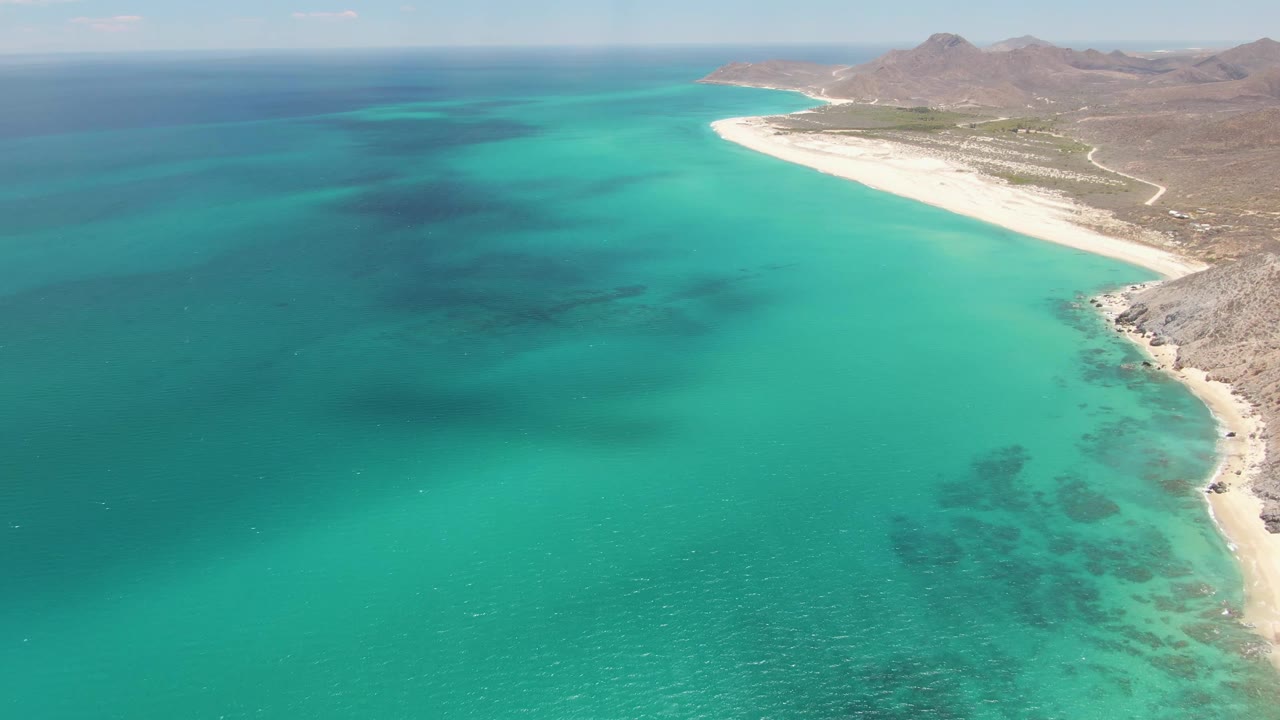 Wide aerial frame at Barakas Beach with the turquoise bay filling the left side, the sandy shoreline sweeping along the right, rocky foreground coast, distant mountains, and no visible people.