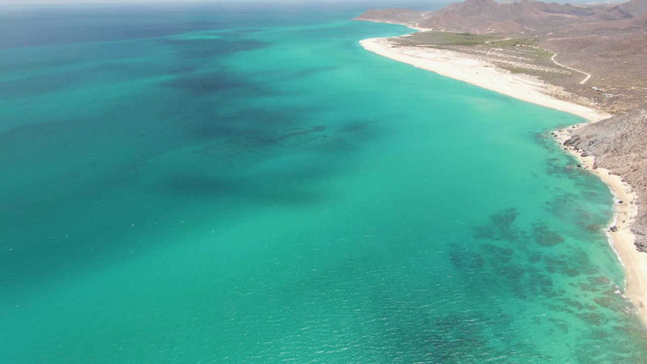 Aerial frame over Barakas Beach with open turquoise water taking most of the view, deeper blue water to the left, a bright sandy beach curving toward rocky land, and no visible people.