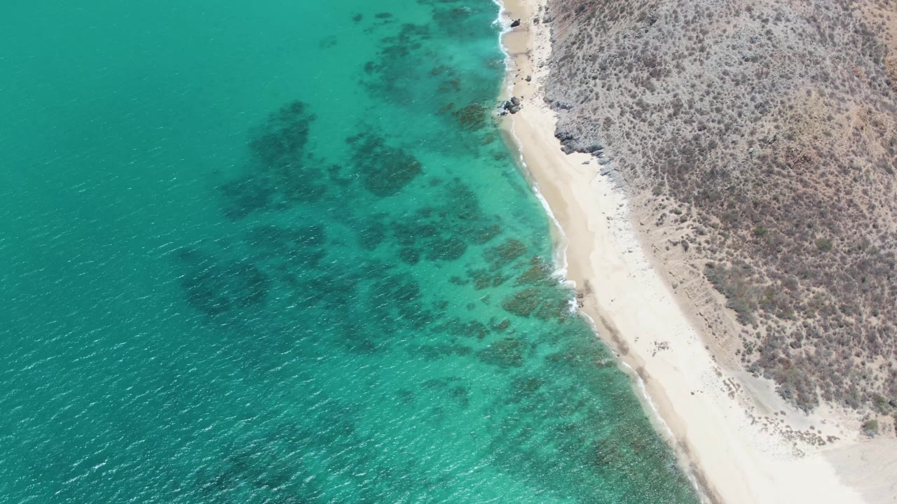 Aerial frame over Barakas Beach in Baja California Sur showing turquoise water, dark underwater patches, a pale curved beach, scrubby rocky land, and tiny beachgoers near the shore.