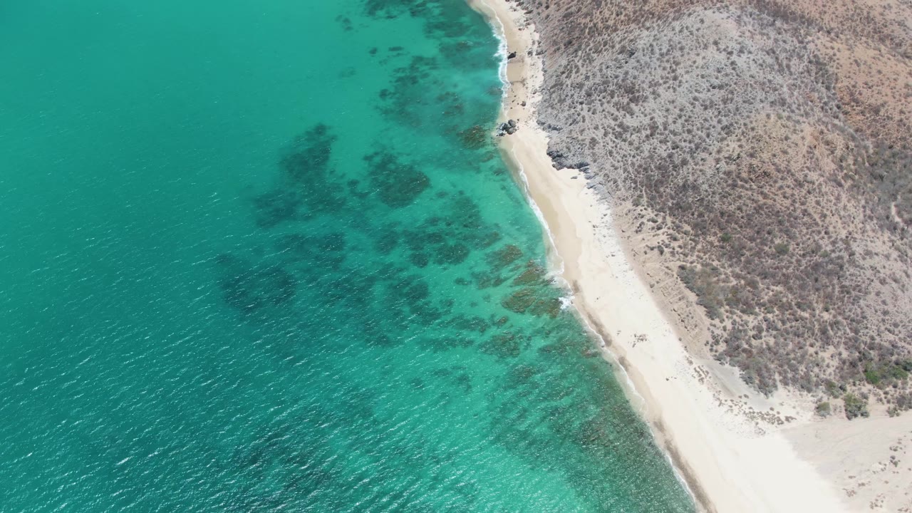 At 0:02, Barakas Beach appears from above with the shoreline running diagonally, deeper teal water to the left, pale sand at the edge, rocky scrubland to the right, and tiny people visible along the beach.