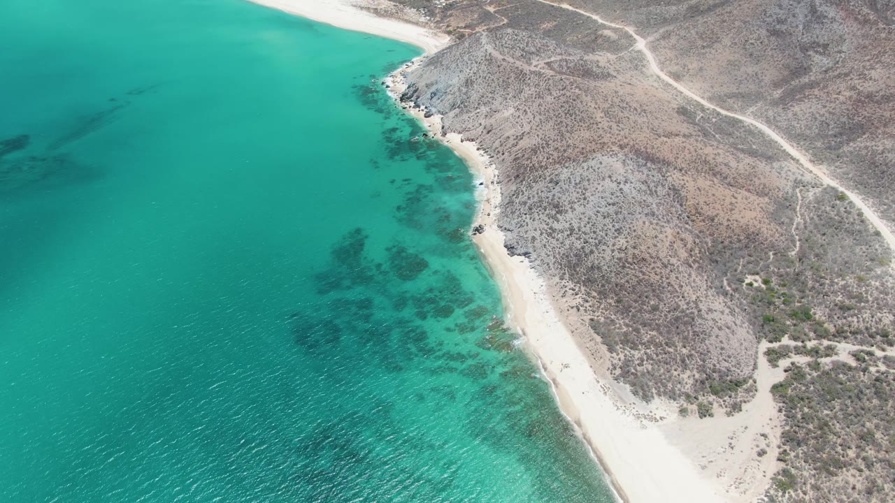Aerial frame over Barakas Beach showing a diagonal sandy shoreline, turquoise shallows with dark underwater patches, rocky land, inland dirt roads, and no visible people.