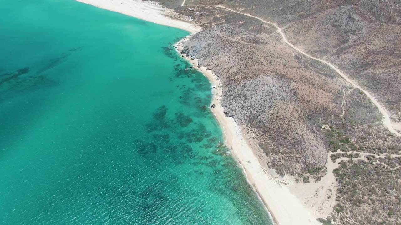 Drone view of Barakas Beach with the shoreline curving along turquoise water, pale sand at the top edge, dry rocky hills inland, dirt tracks, and no visible people.