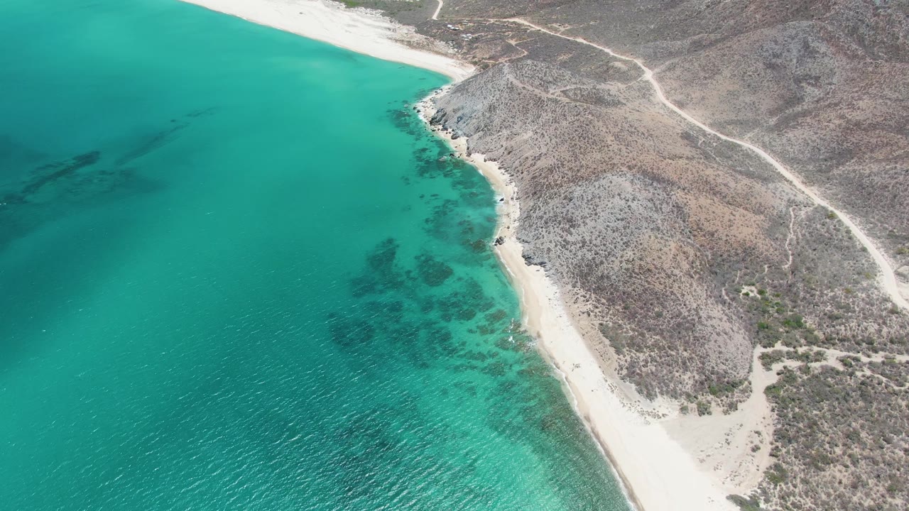 Mid-clip aerial view at Barakas Beach with more open water on the left, a narrow sandy beach through the center, scrub-covered rocky coast to the right, and no visible people.