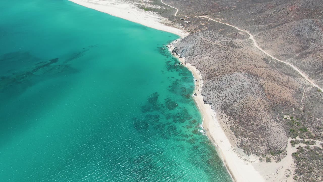 Aerial frame over Barakas Beach in Baja California Sur showing turquoise water on the left, a narrow sandy beach, scrubby rocky hills, dirt tracks, and no visible people.