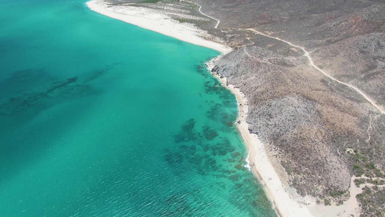 Aerial view at Barakas Beach with the curving sandy shoreline centered between clear turquoise water, dark underwater patches, dry hills, and no visible people.