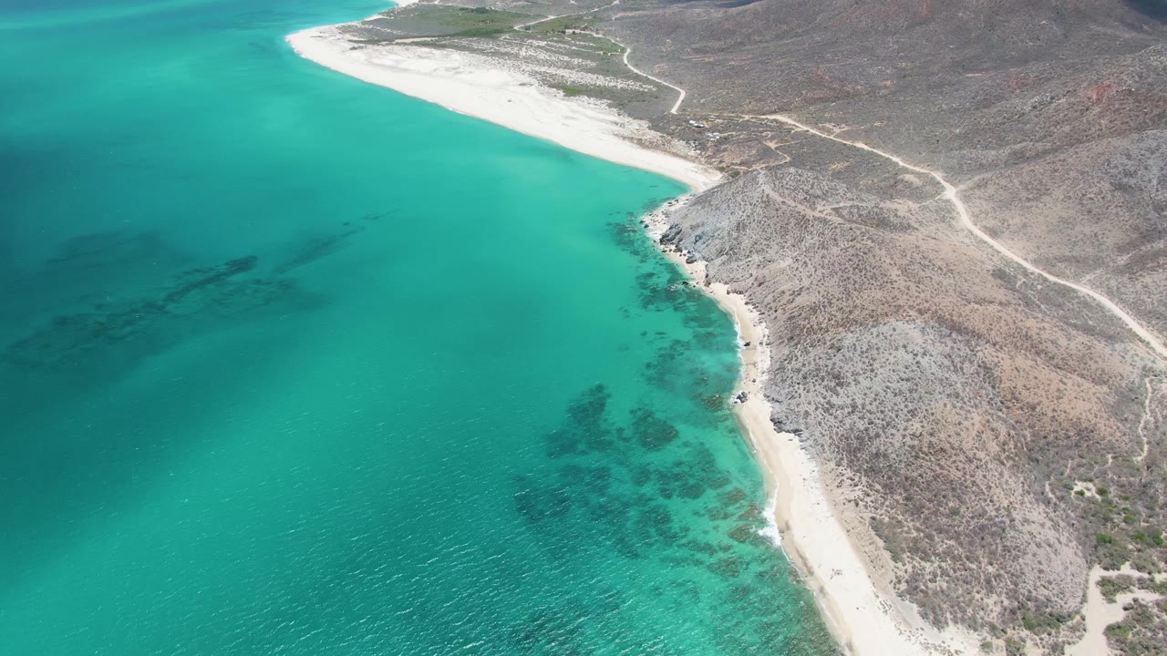 Aerial frame from the Barakas Beach clip showing more of the coastline, with turquoise water spreading left, pale sand along the shore, dry terrain to the right, and no visible people.