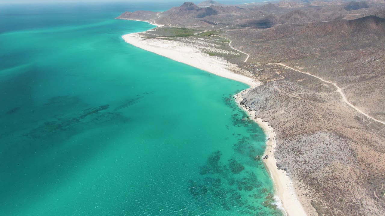 Aerial view of turquoise water beside a white sandy coast and rocky desert hills.