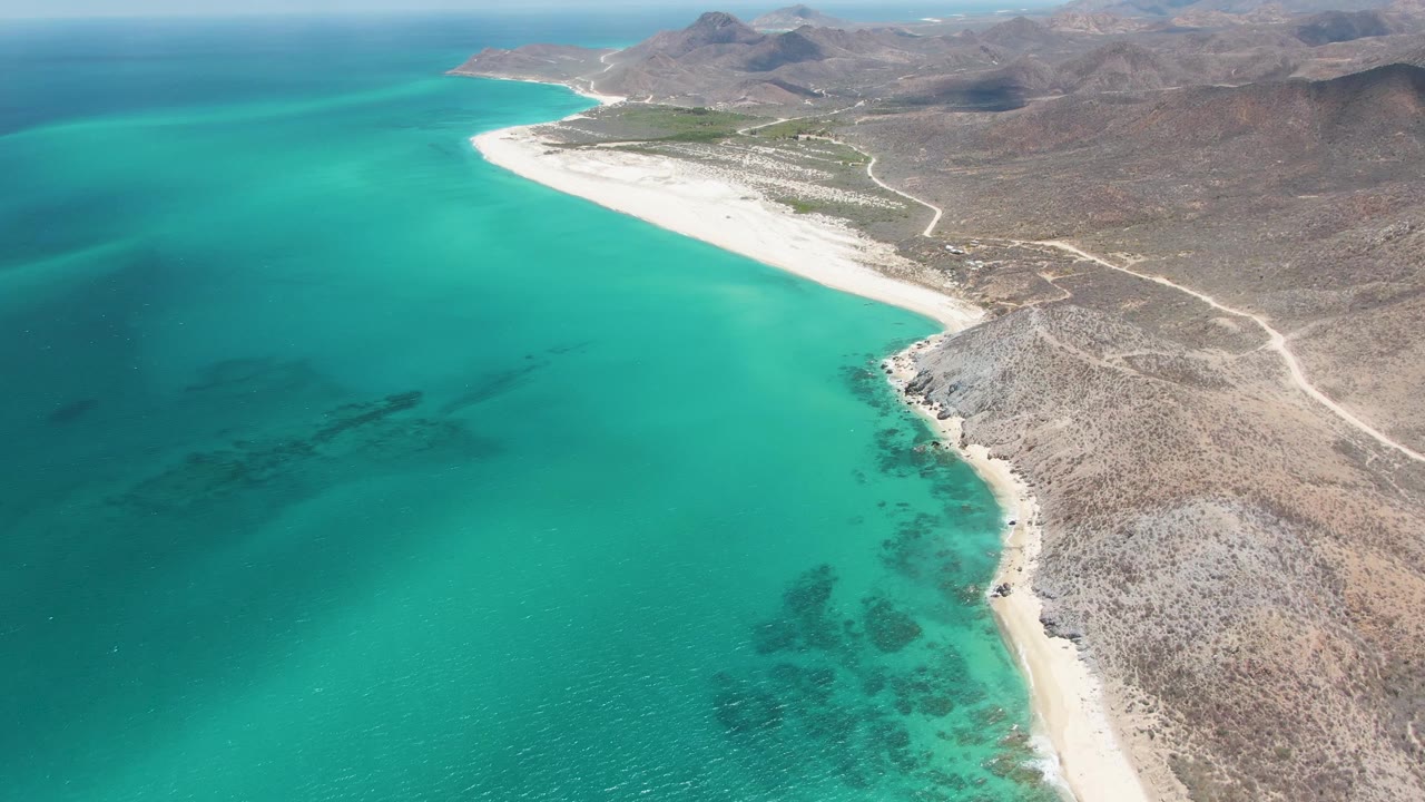 Second aerial moment over Barakas Beach showing more of the curving white sand, clear turquoise sea with darker underwater patches, pale dirt roads across dry land, and no visible people.